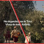 A screenshot from the Airbnb Tour de France 2026 campaign ad featuring the French text "Ne regardez pas le Tour. Vivez-le avec Airbnb" (Don't watch the Tour. Live it with Airbnb.) over a scenic view of an olive grove and a family walking on a path, with the Airbnb logo in the top right corner.